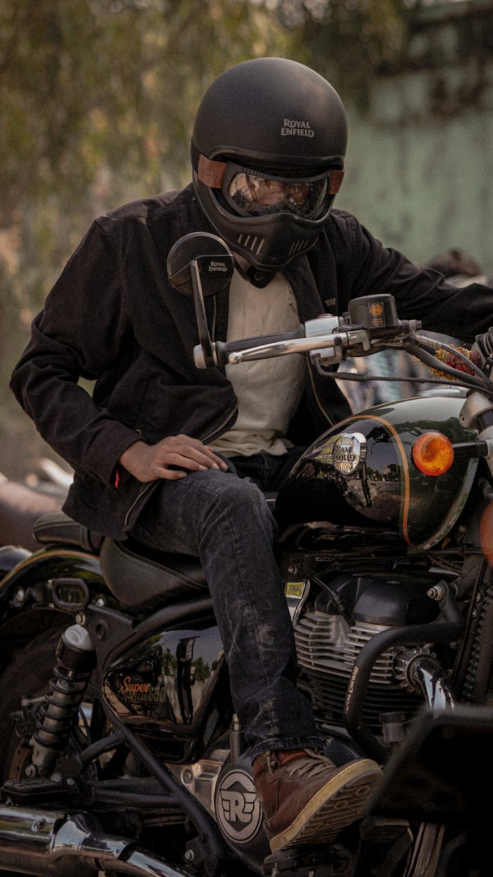 A biker in a helmet and black jacket rides a Royal Enfield motorcycle on a sunny day.